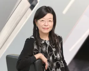 Asian woman with glasses wearing a black dress with a white floral pattern, sitting on a black chair against a modern background.