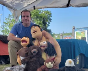 A person sits with a selection of puppets surrounding him
