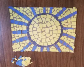 A nearly-complete sun mosaic sits on a table. A dark blue ring and royal blue rays represent the sun. The centre of the sun is filled with yellow. Unused blue and yellow squares sit in a pile next to the mosaic design