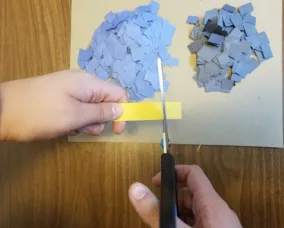 Des mains tenant des ciseaux découpent une bande de papier jaune en petits carrés sur une table. Des piles de carrés bleus et noirs prédécoupés sont posées sur une feuille de papier.