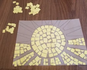 A sun design on cardboard sits on a table. The centre of the sun and half of the space between sun rays are filled with yellow squares. Additional piles of yellow squares sit on the table next to the design.