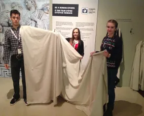 Une extrémité du morceau de tissu blanc pend sur l'épaule du jeune homme et couvre tout son bras. Les jeunes femmes tiennent le reste du tissu.