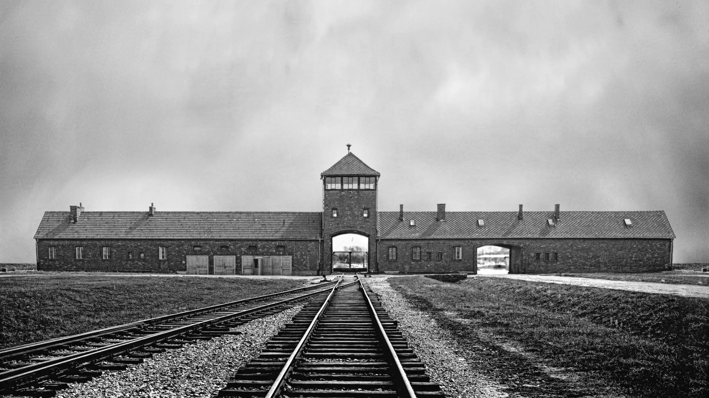 Black and white archival image showing train tracks in the foreground leading into the distance towards the entrance to the Auschwitz camp.