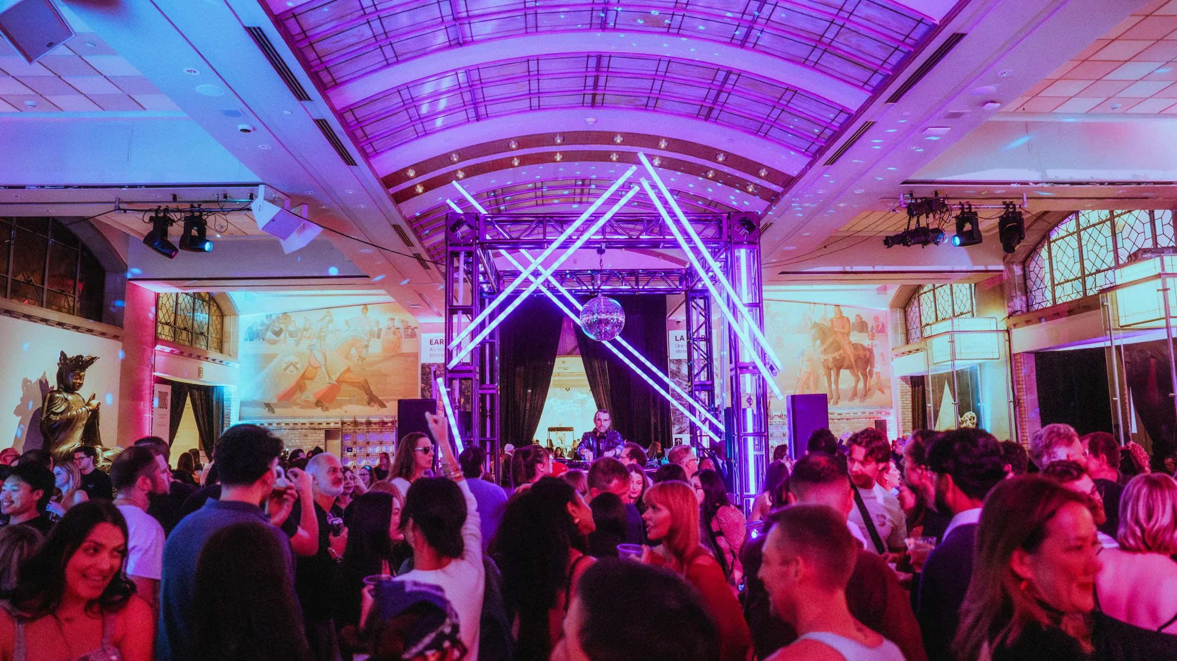 A crowd in a museum conversing. In the background is a DJ under a disco ball.