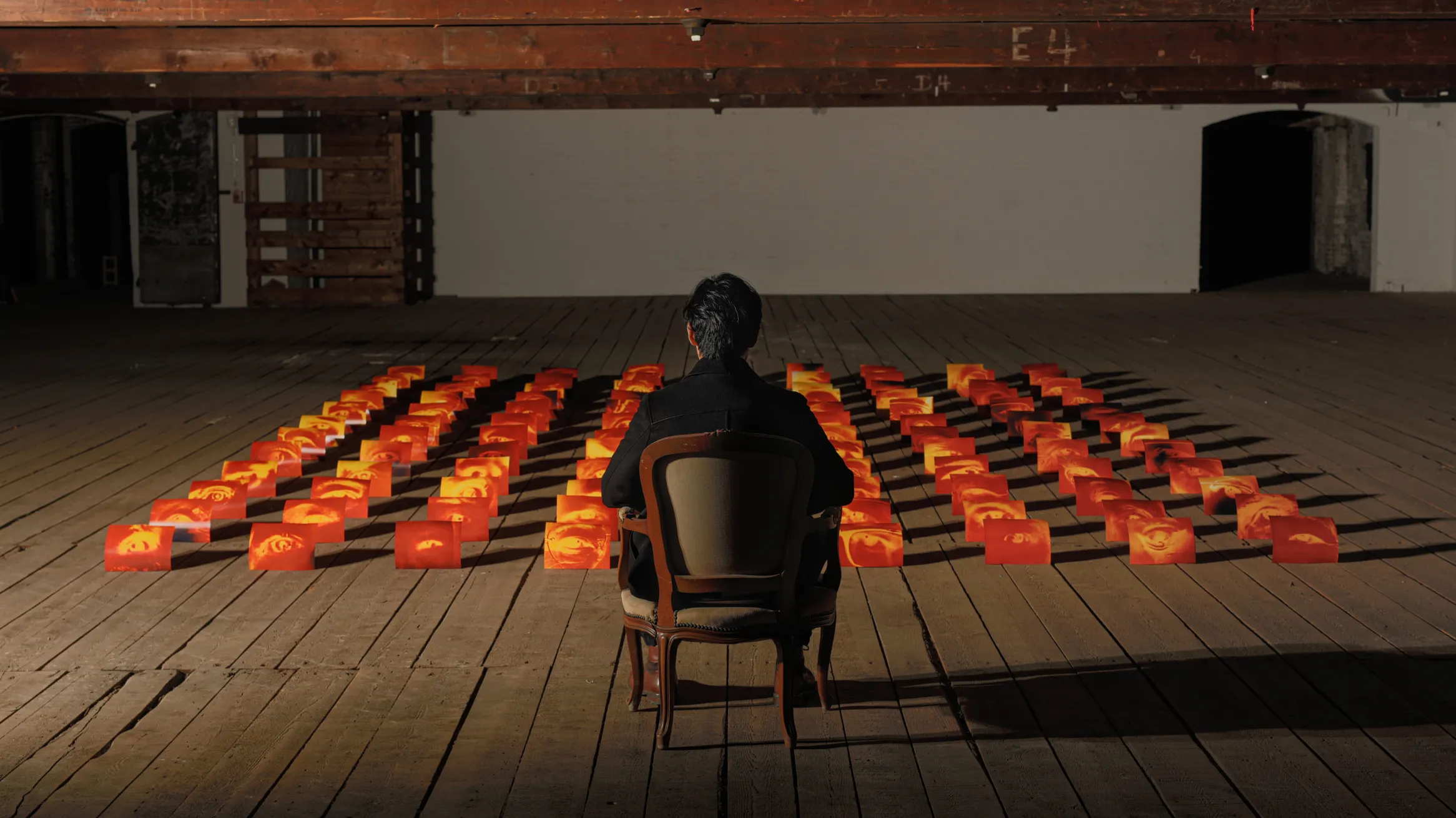 A person sits alone in a wooden chair facing an art installation of glowing orange and red blocks arranged in a grid, each block featuring a human eye, set in a dimly lit industrial-style room with exposed beams and wooden flooring.