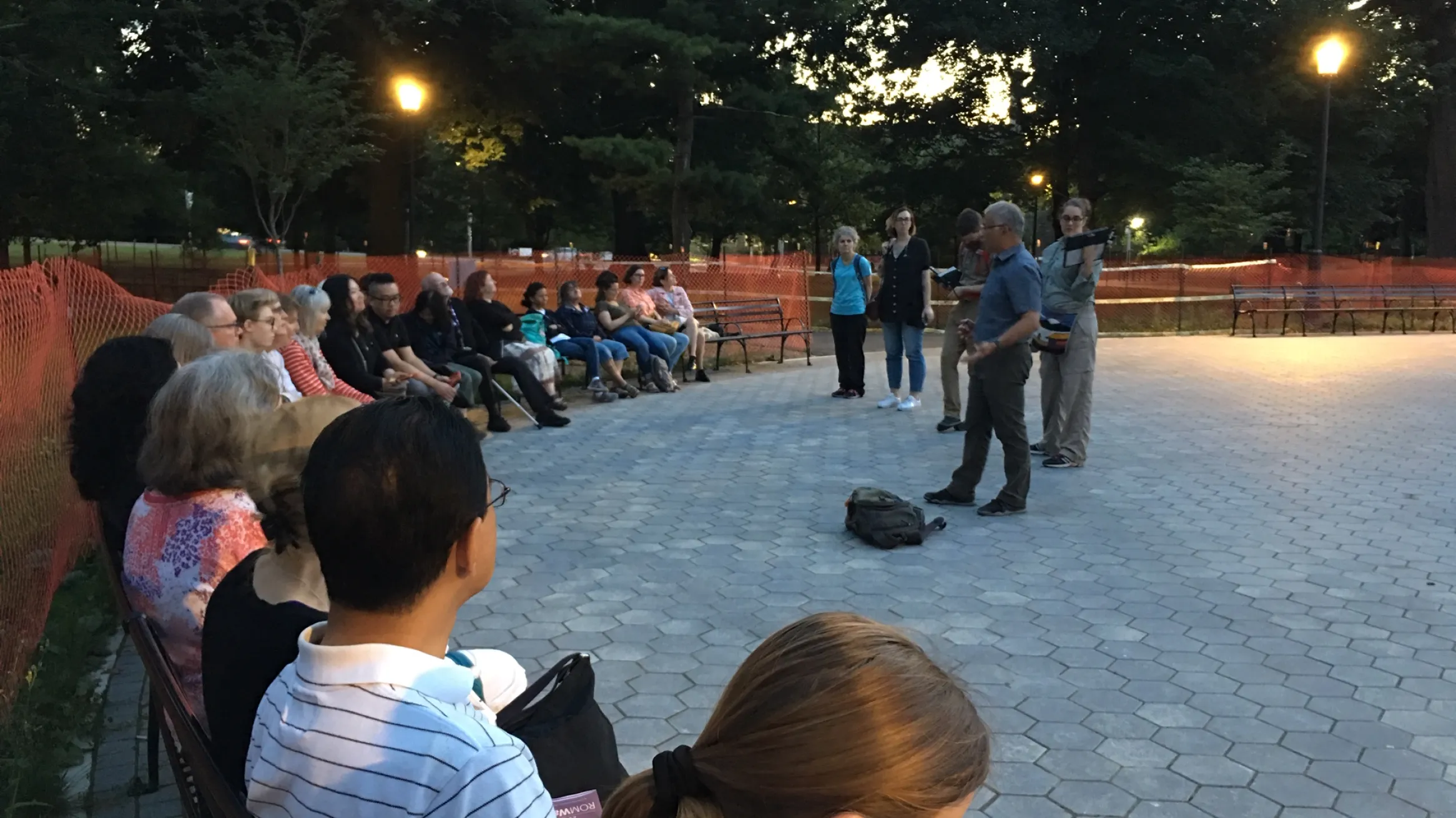Group of people seated in a park at dusk, listening to a speaker.