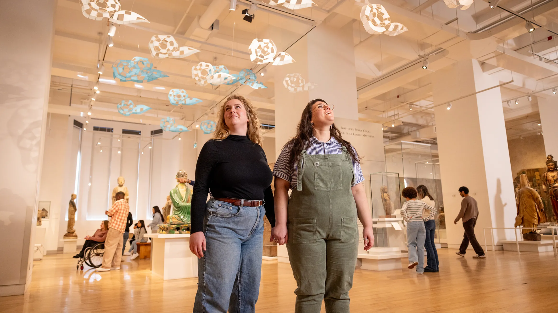 Two visitors hold hands while looking up at a hanging art installation in a museum gallery.