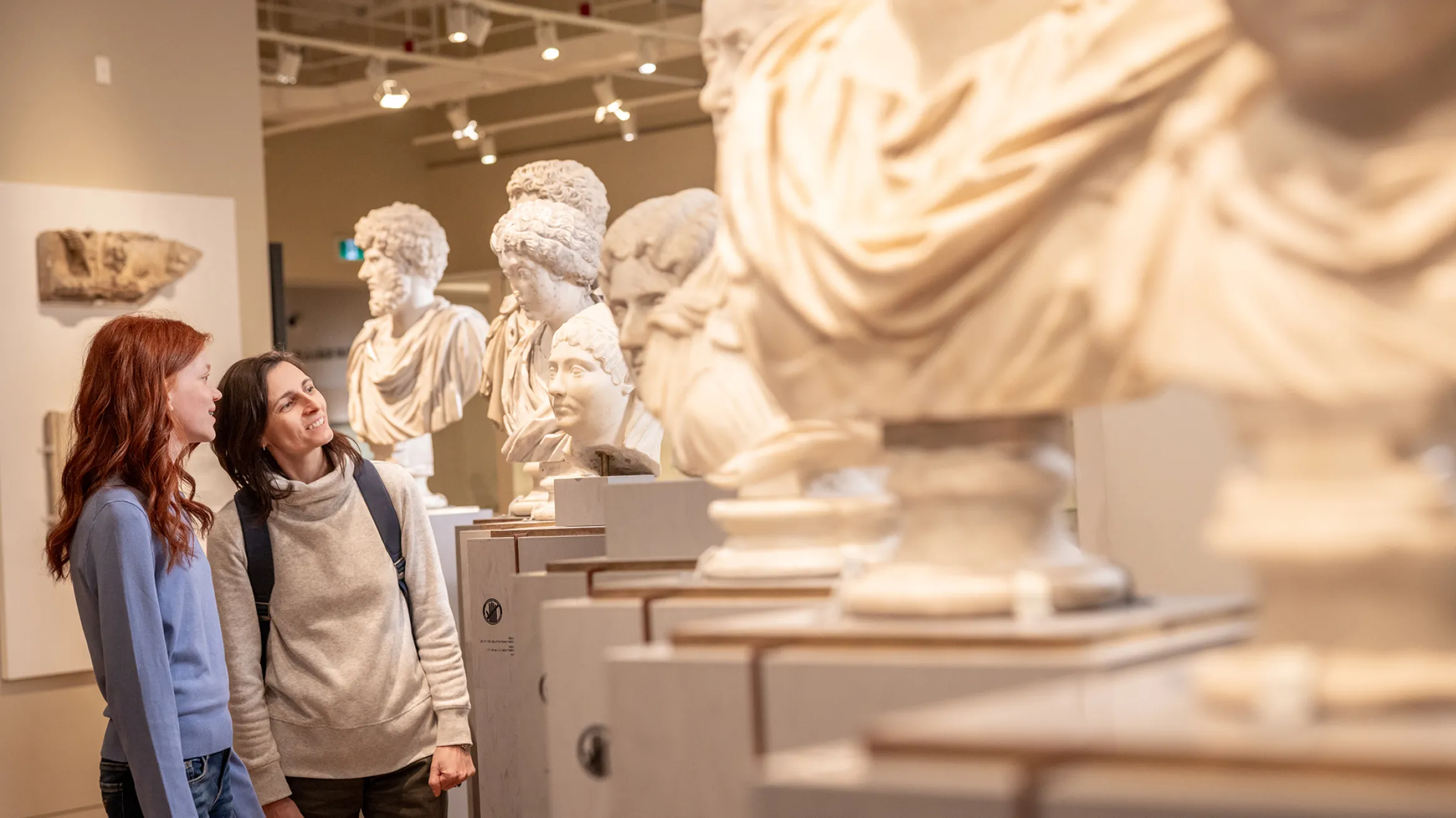 Two visitors look at a row of ancient marble busts on display in a museum gallery.