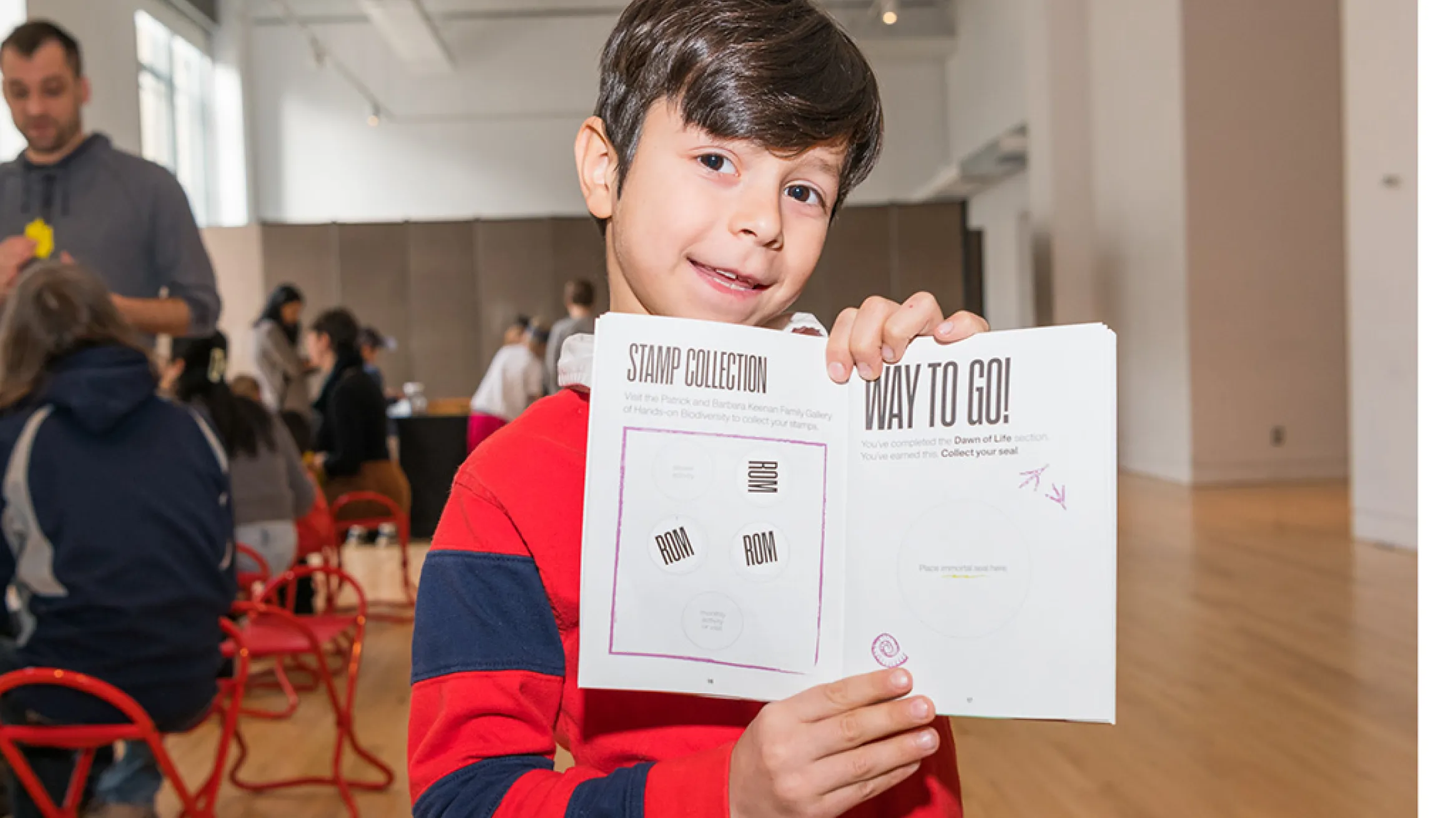 Boy in a red and blue striped sweater holding a white paper booklet with a QR code and text, indoors at an event.
