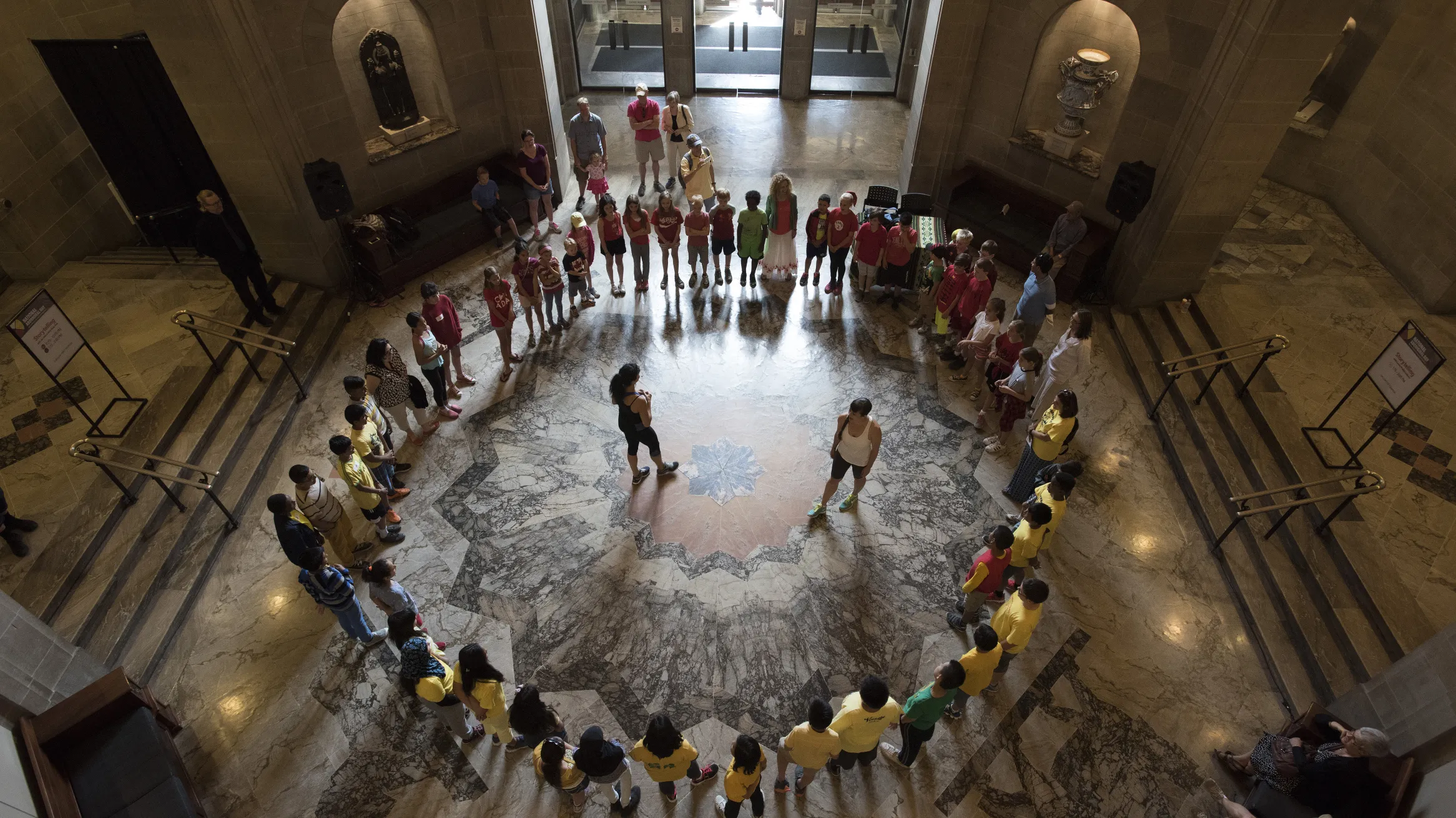The camera looks down from above at group of students sitting in a circle at the bottom of a round ornate room