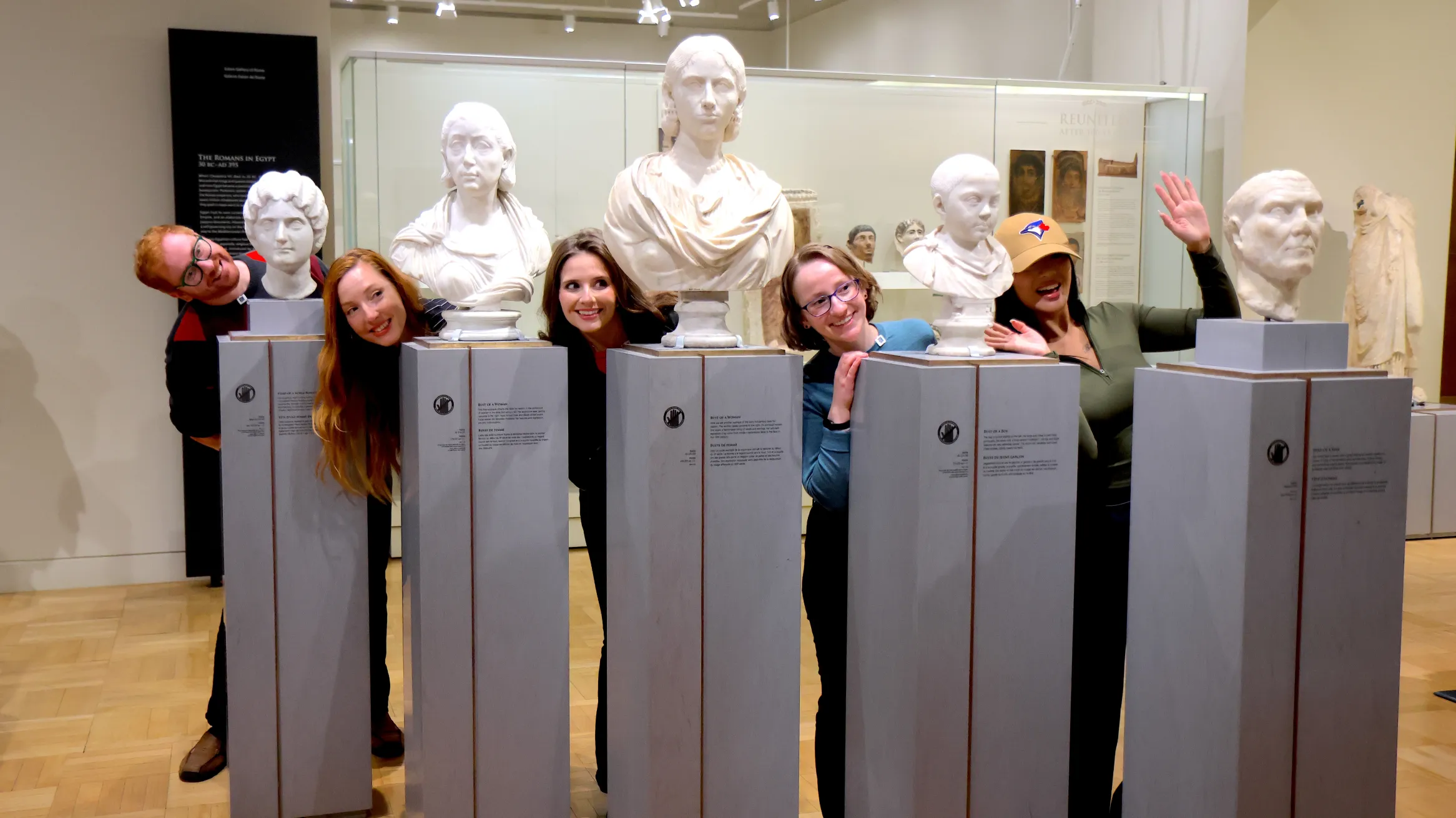 “A group of people playfully pose by leaning out between classical marble busts in a museum gallery.
