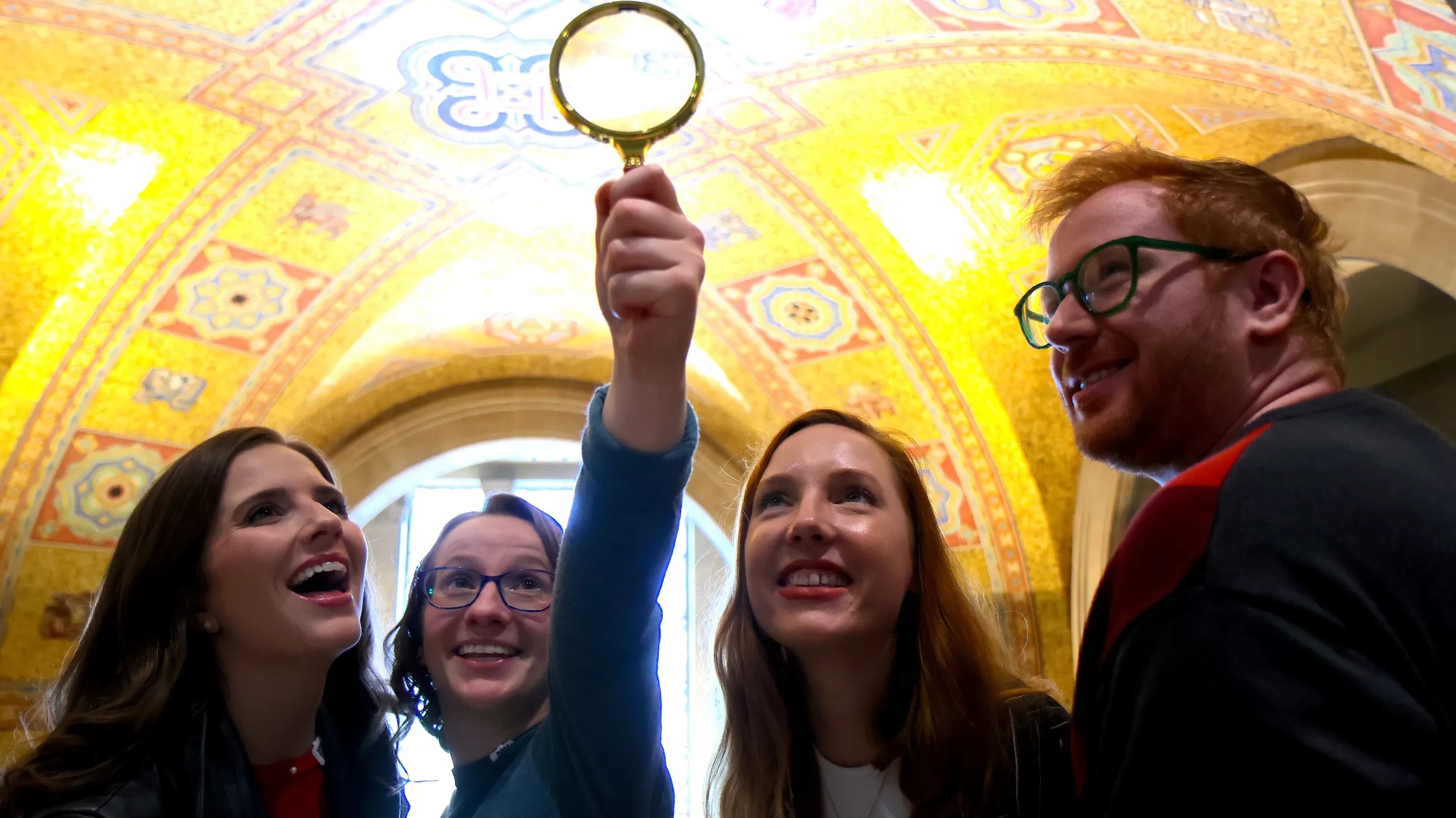 A group of people smile and hold up a magnifying glass while exploring the ornate golden ceiling of the museum rotunda.