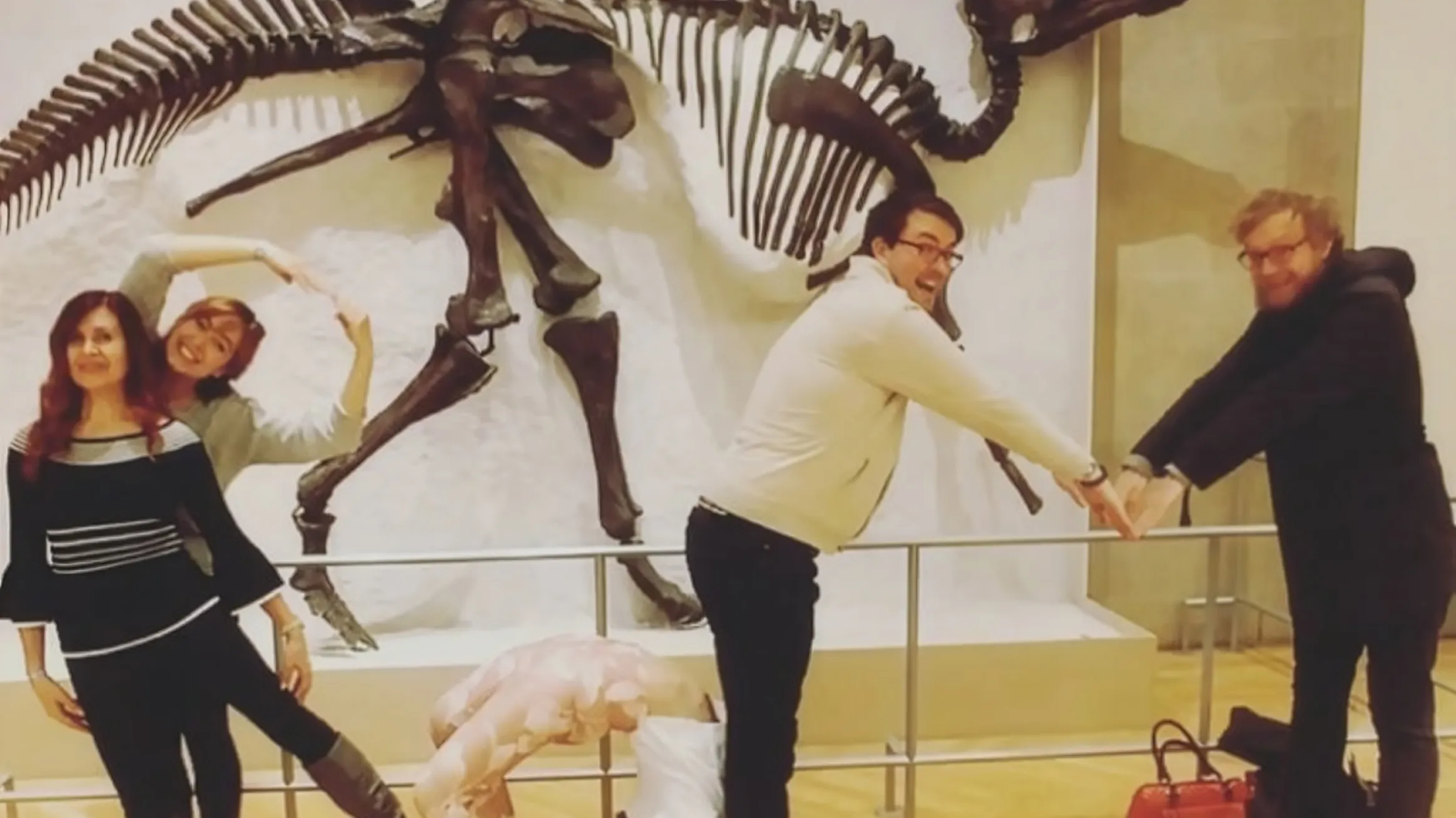 A group of people poses playfully in front of a dinosaur skeleton, arranging their bodies to spell out shapes.