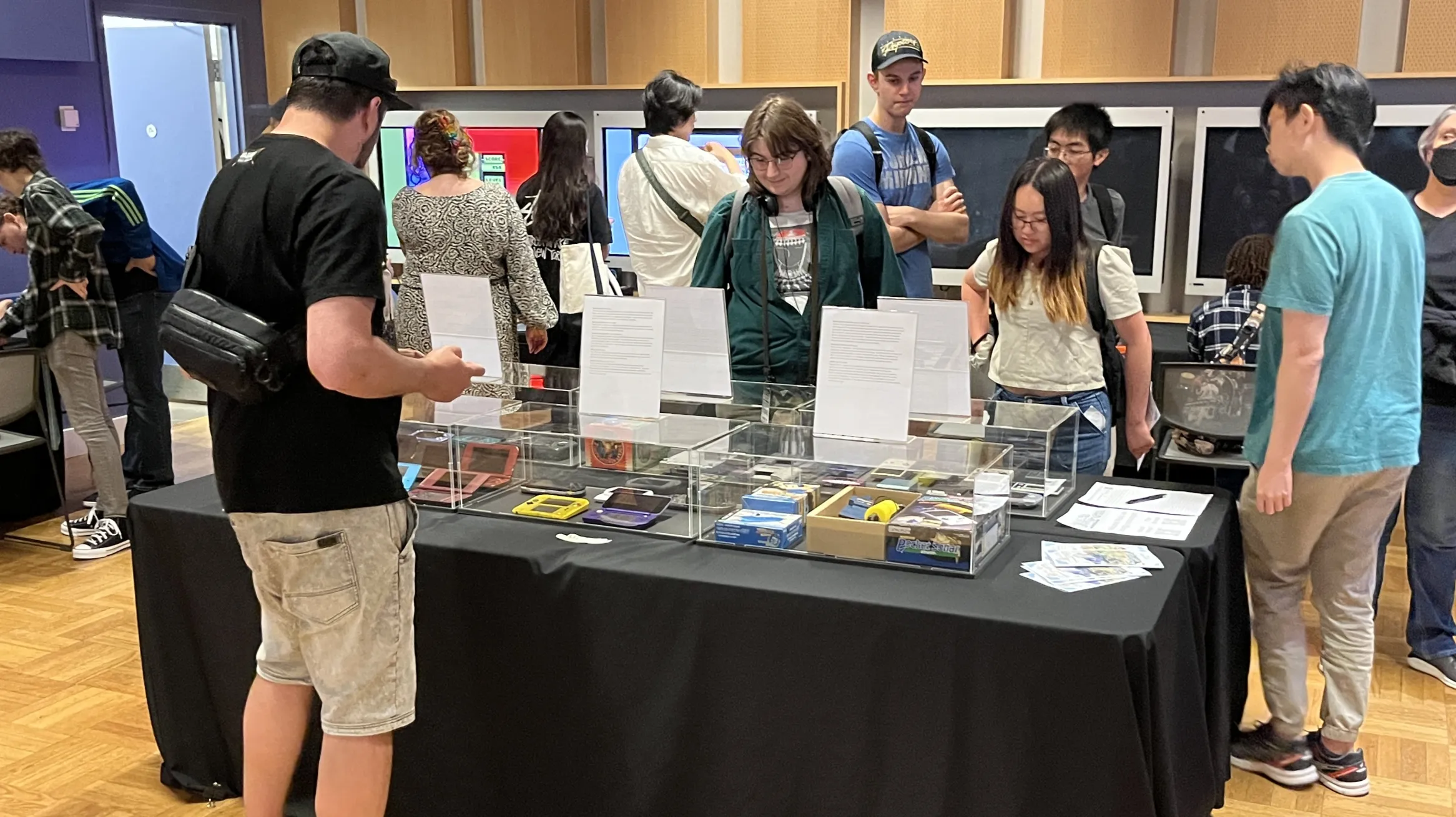 Several people viewing a table covered in a selection of gaming consoles
