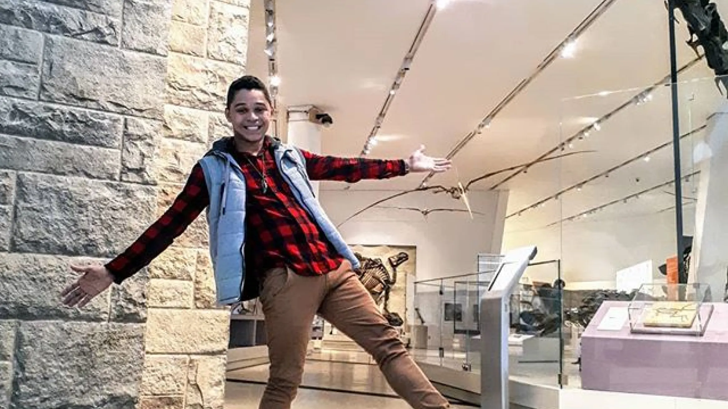 A person smiling and posing with arms outstretched in a museum gallery, standing beneath the long curved neck of a large dinosaur skeleton display.