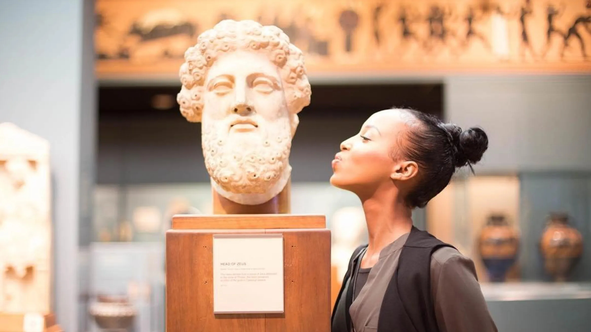 A girl leans toward a large marble bust in a museum as if giving it a kiss, with ancient artifacts and softly lit exhibits visible in the background.