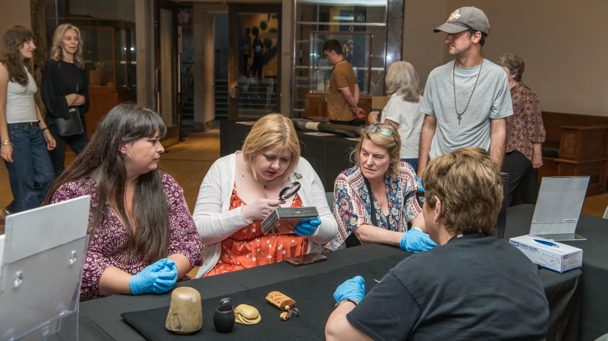 Four people seated at a table examining fossilized skulls, with visitors and a museum entrance in the background.